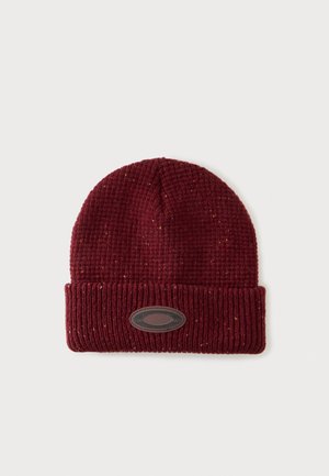 Gorro de punto burdeos con puño doblado y parche ovalado oscuro sobre fondo claro y liso.