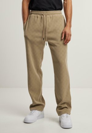 Homme portant un pantalon de jogging beige texturé avec taille à cordon, mains dans les poches, baskets blanches et chemise noire, debout sur un sol blanc.