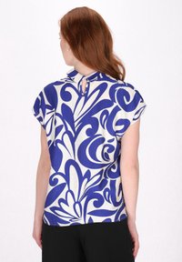 Kurzärmlige Bluse mit einem blau-weißen Blumenmuster, mit Kragen und einem Schlüssellochdetail auf der Rückseite. Hergestellt aus einem leichten Stoff.