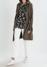 Trench coat leggero verde oliva indossato sopra una camicia nera a stampa floreale, abbinato a pantaloni bianchi a gamba dritta e sneakers bianche.