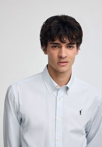 Ljusblå och vit randig skjorta med knappar och krage, med en liten broderad logga på bröstet och en slät textur.