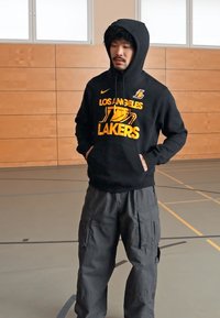 Moletom preto com "LOS ANGELES LAKERS" em amarelo, calças cargo cinzentas, usado numa academia com paredes de madeira e um chão liso.