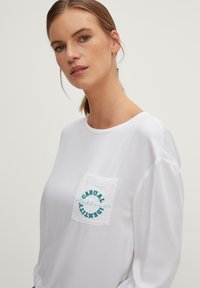 Camicia bianca a maniche lunghe con scollatura rotonda e tasca sul petto. La tasca presenta un ricamo teal con la scritta "CASUAL" e "DENTAL."