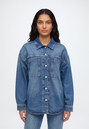Femme aux longs cheveux foncés portant une veste en denim bleue boutonnée avec de grandes poches avant et un jean bleu assorti sur un fond uni.