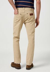 Beżowe spodnie proste z dwoma tylnymi kieszeniami i skórzaną łatą. Materiał ma gładką fakturę i wzmocnione szwy.
