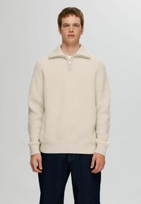 Suéter de cuello redondo y con cremallera en color blanco roto, de textura acanalada, con mangas largas y dobladillo ajustado, que se lleva sobre unos pantalones vaqueros oscuros.