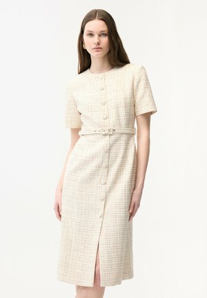 Vrouw draagt een lichtgekleurde midi-jurk met korte mouwen en knopen, voorzien van een subtiel ruitpatroon en een bijpassende riem in de taille.