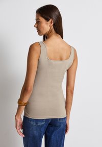 Top sin mangas color beige con escote cuadrado y tirantes anchos, hecho de una tela suave. Combinado con jeans de mezclilla azul y una pulsera ámbar.