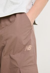 Main dans la poche de shorts cargo mauves avec le logo New Balance visible et une partie d'une chemise gris clair au-dessus des shorts.