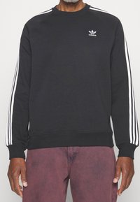 Person som bär en svart Adidas-tröja med vita tre-rands detaljer på ärmarna och vinröda byxor, visad från halsen till höfterna.