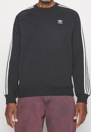 Persoon wearing een zwart Adidas sweatshirt met witte drie-streep details op de mouwen en bordeauxrode broek, weergegeven van nek tot heupen.