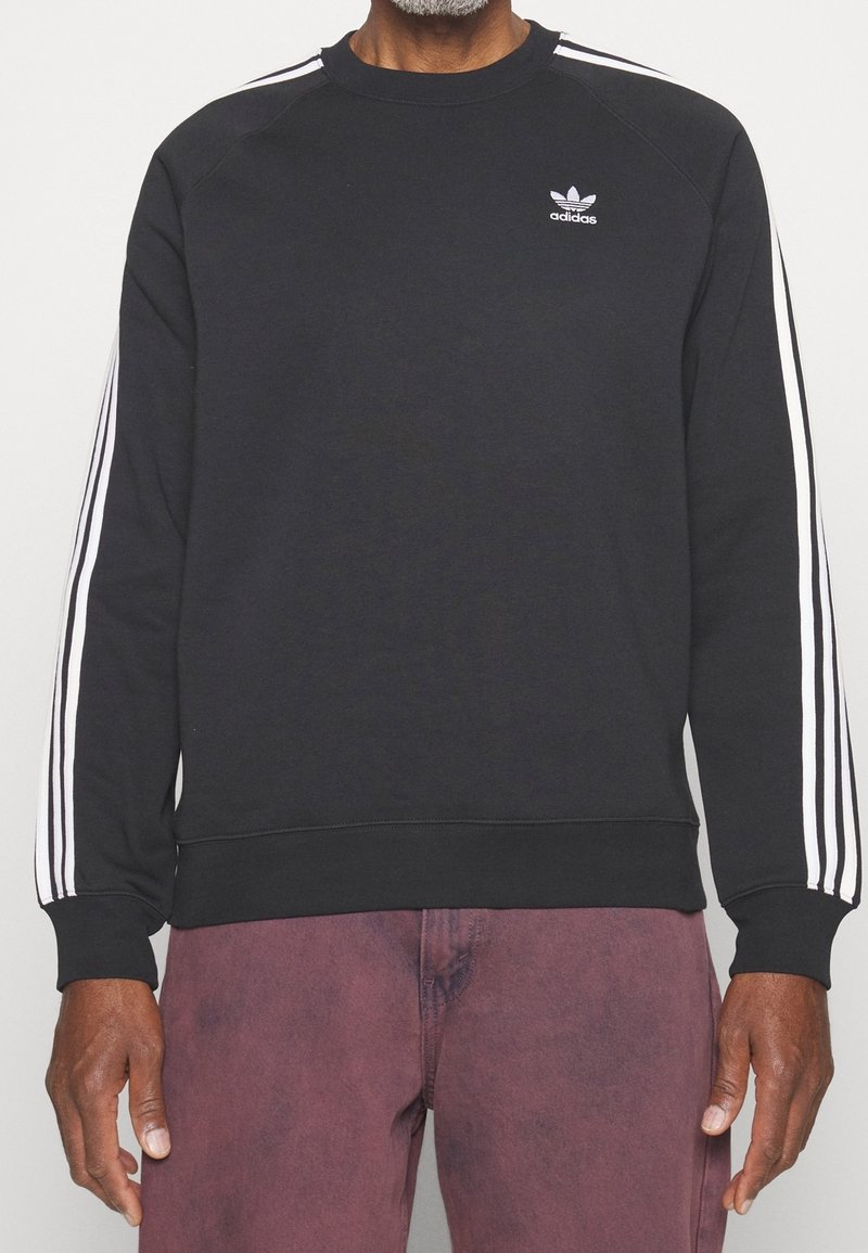 Person som bär en svart Adidas-tröja med vita tre-rands detaljer på ärmarna och vinröda byxor, visad från halsen till höfterna.