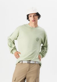 Chemise à manches longues vert clair avec des imprimés graphiques sur les manches, associée à un pantalon cargo beige et une casquette blanche. Tissu doux, coupe décontractée.