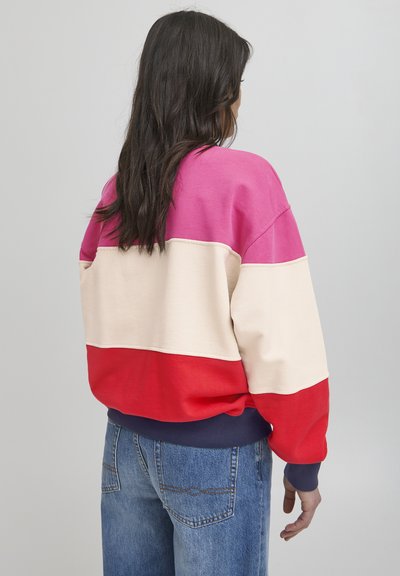 Krāsainais svīteris rozā, krēmkrāsā un sarkanā tonī ar ribotu tumši zila kaklu. Audums no kokvilnas maisījuma; brīvais griezums ar kritušām plecu šuvēm.