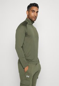 Olíva zöld, hosszú ujjú, cipzáras felső, könnyű anyagból készült, magas gallérral és testhezálló fazonnal. Az Under Armour logó a mellkasán.