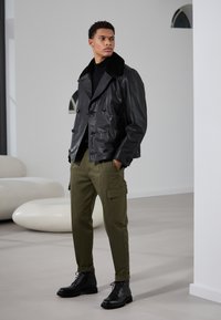 Veste double boutonnage en cuir noir avec un col en fausse fourrure, pantalon cargo vert olive et bottes noires à lacets, le tout sur un fond minimaliste.
