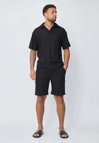 Chemise noire à manches courtes avec un col, associée à un short noir, fabriqué en tissu léger. Porté avec des sandales à glisser noires.