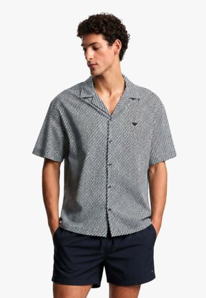 Giovane uomo con capelli ricci che indossa una camicia a maniche corte con bottoni e pantaloncini neri, mani in tasca, guarda di lato.
