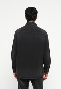 Chemise en denim noir à manches longues avec un col boutonné, un ourlet courbé et des poignets à un bouton, présentant une texture lisse et des couleurs atténuées.