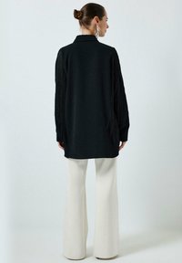 Long black shirt with fringed sleeves, paired with white flared trousers. The fabric appears soft, with a smooth texture and collar detail.