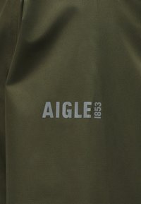 Ύφασμα ελιάς με ανοιχτό γκρι τυπωμένο κείμενο "AIGLE 1853", που δείχνει υφή και ελαφριές πτυχώσεις.