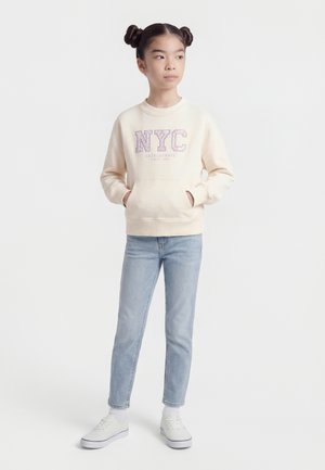 Mladé dievča s účesom s dvojitým drdolom, oblečené v krémovej mikine NYC, svetlomodrých džínsoch a bielych teniskách, stojace s rukami v vreckách.