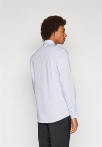 Hellblaues, langärmliges Hemd mit Kragen, Rückennähten und Knopfmanschetten. Der Stoff scheint strukturiert und leicht zu sein. Getragen mit dunklen Hosen.