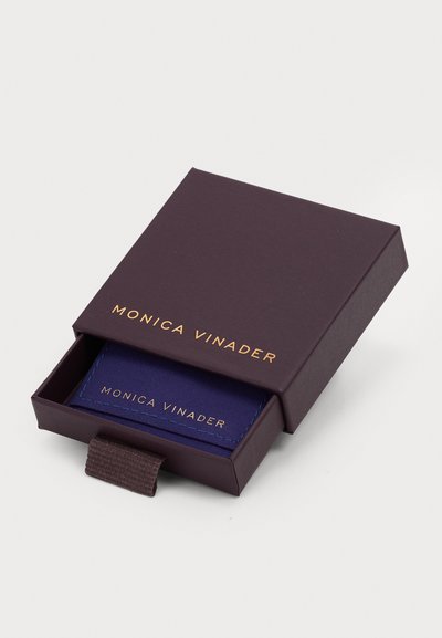 Lila doboz arany színű felirattal "MONICA VINADER", félig nyitva, belsejében egy sötétkék kártyával, kék varrással és hasonló márkázással.