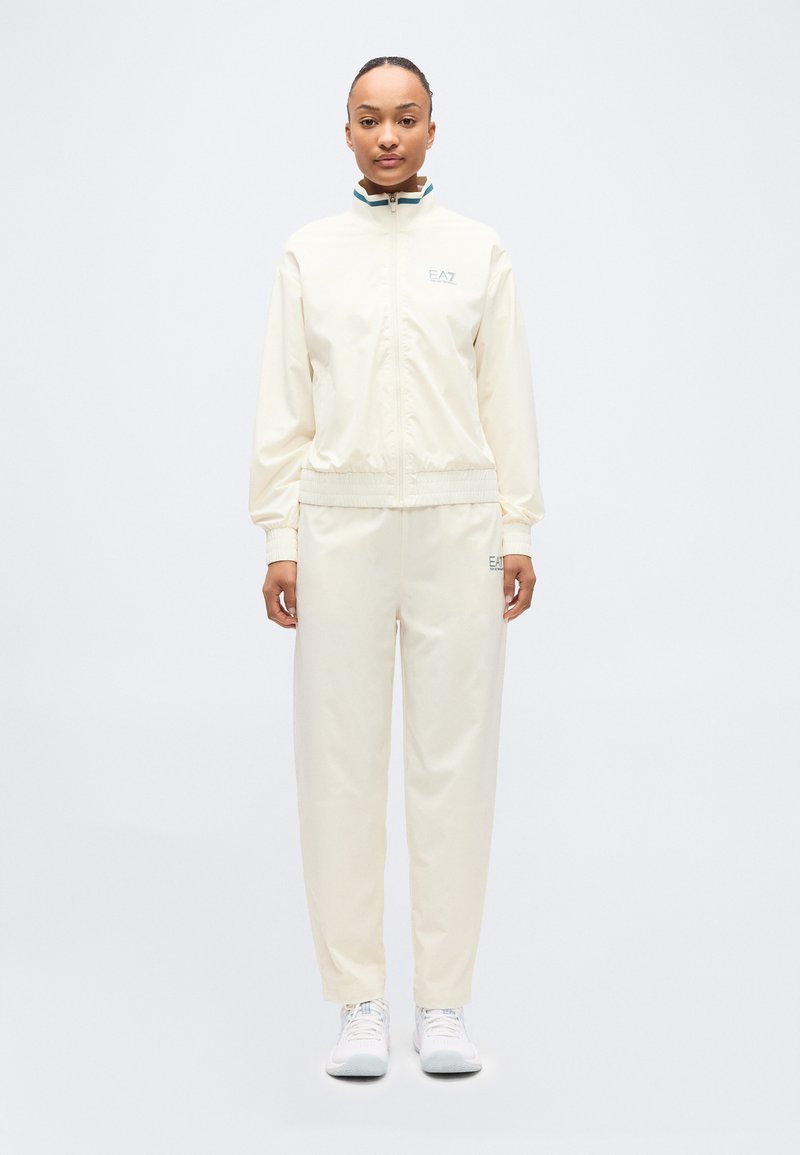 Γυναίκα στέκεται με το πρόσωπο προς τα εμπρός, φορώντας μια κρεμ φόρμα με το λογότυπο EA7 και λευκά αθλητικά παπούτσια, μπροστά σε απλό λευκό φόντο.