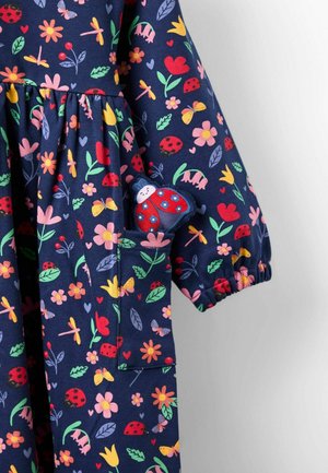 Tengeri kék ruha színes virág- és rovarmintával. Rugalmas mandzsettákkal, ráncolt derékkal és egy zsebből előbukkanó katicabogár plüssjátékkal rendelkezik.