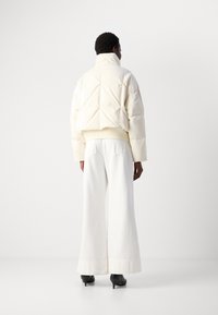 Persoon in een crèmekleurige pufferjack, met wijde witte broek en zwarte hakken, staand tegen een effen witte achtergrond.