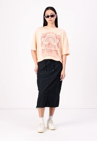 Ferskenfarvet oversized t-shirt med et grafisk print, parret med en sort midi-nederdel. Modellen har hvide sneakers og mørke solbriller på.
