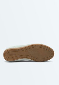 Semelle d'une sneaker de couleur claire avec une semelle extérieure en caoutchouc brun à motifs et le logo "OYSHO" près du talon, sur fond blanc.