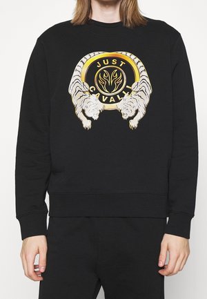 Czarna bluza z okrągłym logo w kolorze złota i czerni, po bokach wspierana przez dwa białe tygrysy oraz napis "Just Cavalli" na piersi.