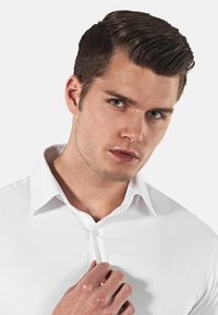 Chemise blanche avec un devant à boutons et un col pointu. Le tissu semble lisse et légèrement ajusté, mettant en valeur le design structuré.
