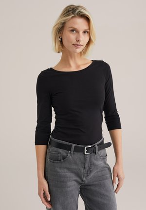 Haut noir à manches longues avec un large décolleté, associé à un jean gris taille mi-haute et une ceinture noire. Le tissu semble doux et ajusté.
