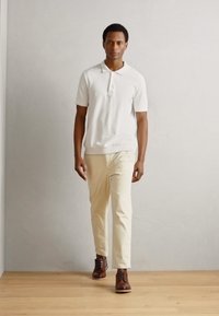 Tricou polo alb cu mâneci scurte, pantaloni crem și pantofi cu șireturi maro. Design simplu cu o croială obișnuită, plasat pe un fundal simplu.
