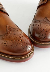 Pantofi din piele maro cu detalii brogue, având perforații, vârf rotund și cusături contrastante pe talpă.