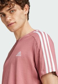 T-shirt in cotone rosa con design a tre strisce bianche sulle spalle e un piccolo logo bianco sul petto, scollatura rotonda e maniche corte.