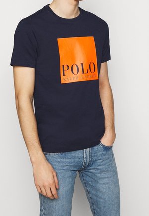 Námořnická modrá bavlněná tričko s lesklým oranžovým čtvercem, na kterém je tučně černým písmem napsáno "POLO" a "RALPH LAUREN".