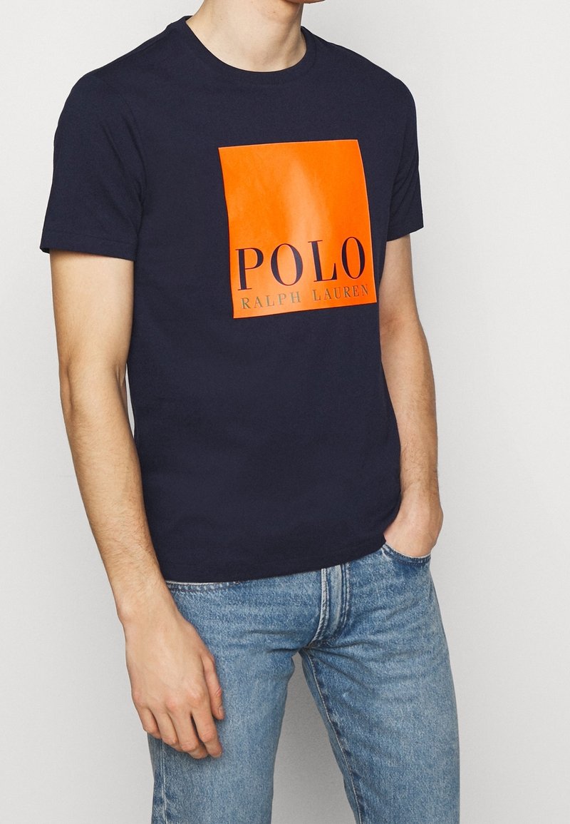 Marinblå bomullströja med en glänsande orange ruta med "POLO" och "RALPH LAUREN" i fet svart text på framsidan.
