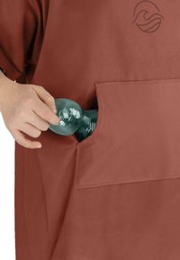 Rostfarbenes Kleidungsstück mit einer großen Tasche, in die eine Hand eine türkisfarbene Plastikflasche einfügt. Glatte, matte Textur mit einem schlichten Logo.
