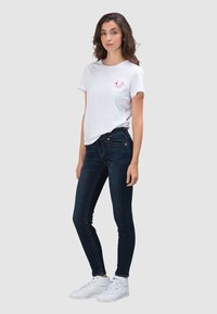 Wit katoenen T-shirt met korte mouwen en een kleine rode afbeelding op de borst, gecombineerd met donkerblauwe skinny jeans en witte hoge sneakers.