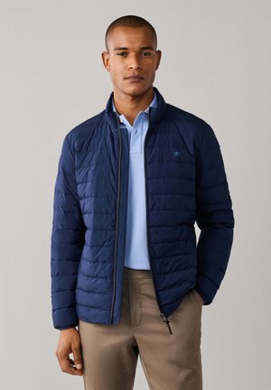 Man wearing a navy quilted jacket over a light blue polo shirt and beige pants, standing against a plain light gray background.