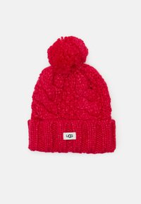 Červená pletená čiapka s pom-pomom na vrchu, s dizajnom káblového pletenia a rebrovým manžetovým úpletom. Na prednej strane je viditeľná štítok s logom UGG.