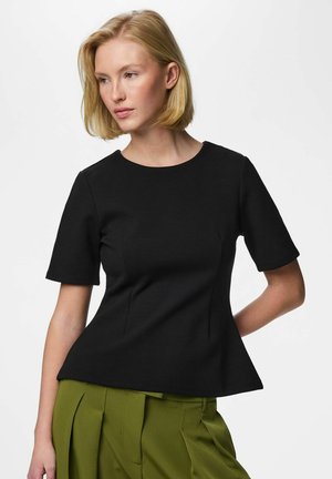 Donna bionda che indossa un top nero aderente a maniche corte e pantaloni plissettati a vita alta color verde oliva, che guarda di lato.