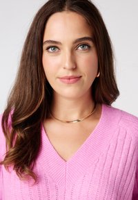 Roze gebreide trui met V-hals en geribbelde textuur, gedragen met een gouden soepele chokersieraad. Het haar is lang en golvend, dat het gezicht omlijst.