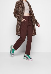 Leopardmönstrad lång kappa, beige kort topp, bruna smalbyxor och färgglada sneakers med gröna accenter och texturerad detaljer.