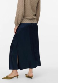 Gonna maxi in satin blu navy con uno spacco laterale, texture liscia e design elegante. Abbinata a un top beige con maniche a palloncino e tacchi kitten verde oliva.