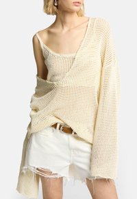 Beige offener Strickpullover mit lockerer Passform und langen Ärmeln, über einem hellen Tanktop getragen. Kombiniert mit ausgefransten weißen Jeansshorts und einem beigen Gürtel.
